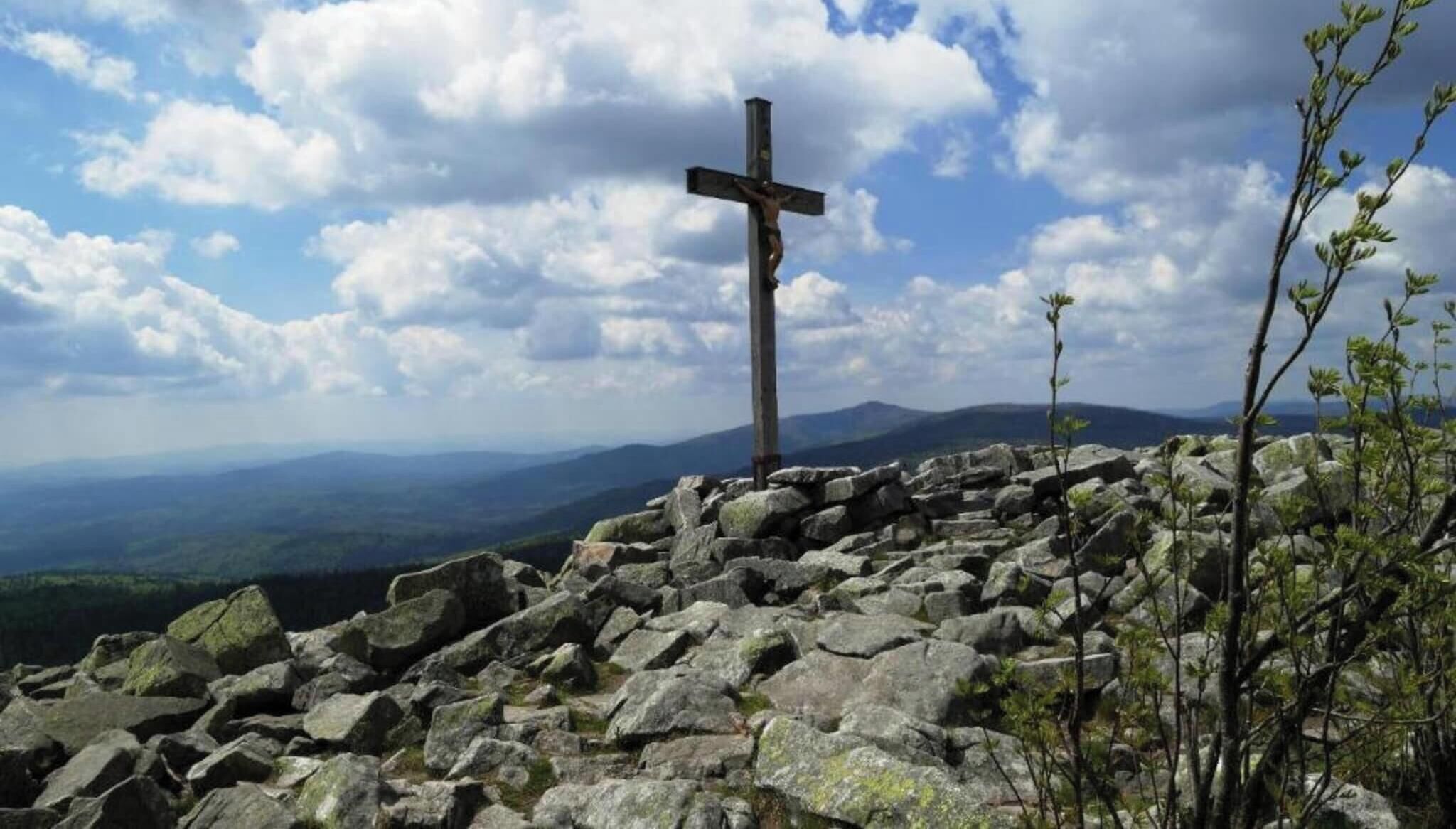 Ein Kruzifix auf einer felsigen Klippe, im Hintergrund eine gebirgige Landschaft.