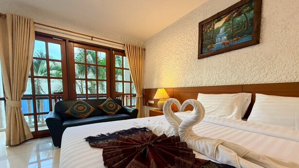 A hotel room with a bed, a sofa, a painting, and a view of palm trees. - Blue Neptuna Hotel Patong (Patong)