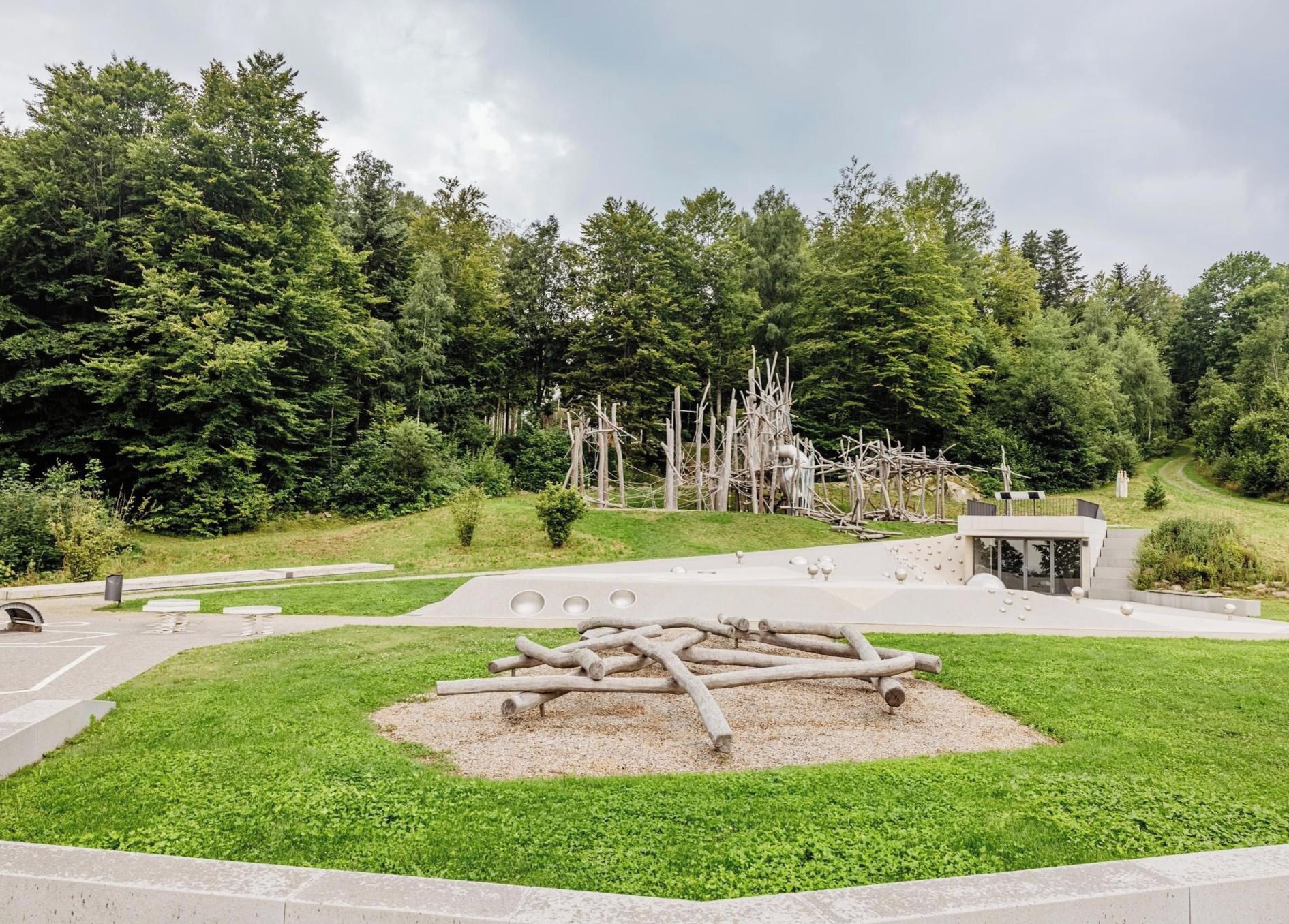 Un parco con un edificio moderno, un'area con ghiaia e tronchi, e un sentiero.