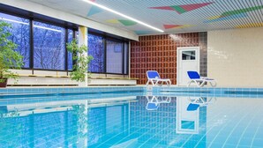 Ein beheiztes Hallenbecken mit klarem, blauem Wasser, zwei blaue Liegestühle und große Fenster mit Blick auf Bäume.