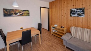 Ein Zimmer mit Holzboden, einem Esstisch mit Stühlen, einer Couch, einer weißen Pendelleuchte und zwei Bildern an den Wänden.