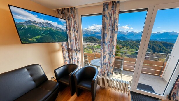 Een kamer met een zwarte leren bank, twee zwarte fauteuils, een aan de muur bevestigde televisie en een balkon met uitzicht op bergen en een meer.