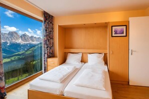 Ein Schlafzimmer mit zwei Betten, einem Holzkopfende, einem großen Fenster mit Bergblick, einem gerahmten Bild an der Wand und einer Tür.