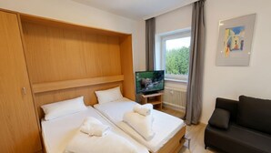 Habitación de hotel con dos camas, televisión, sofá y ventana con vista a árboles.