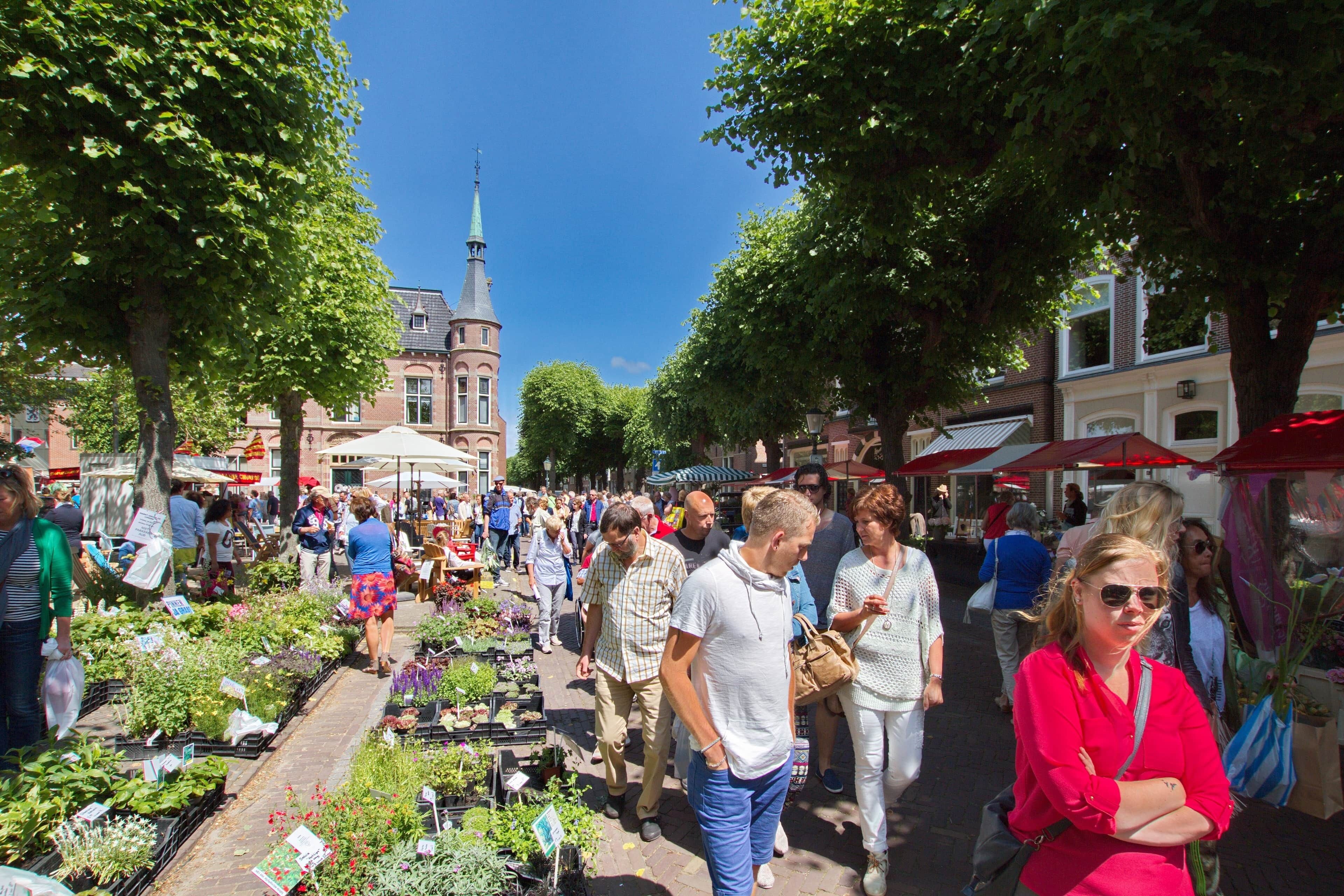 Un mercado al aire libre concurrido, con gente comprando y caminando por una calle bordeada de árboles.