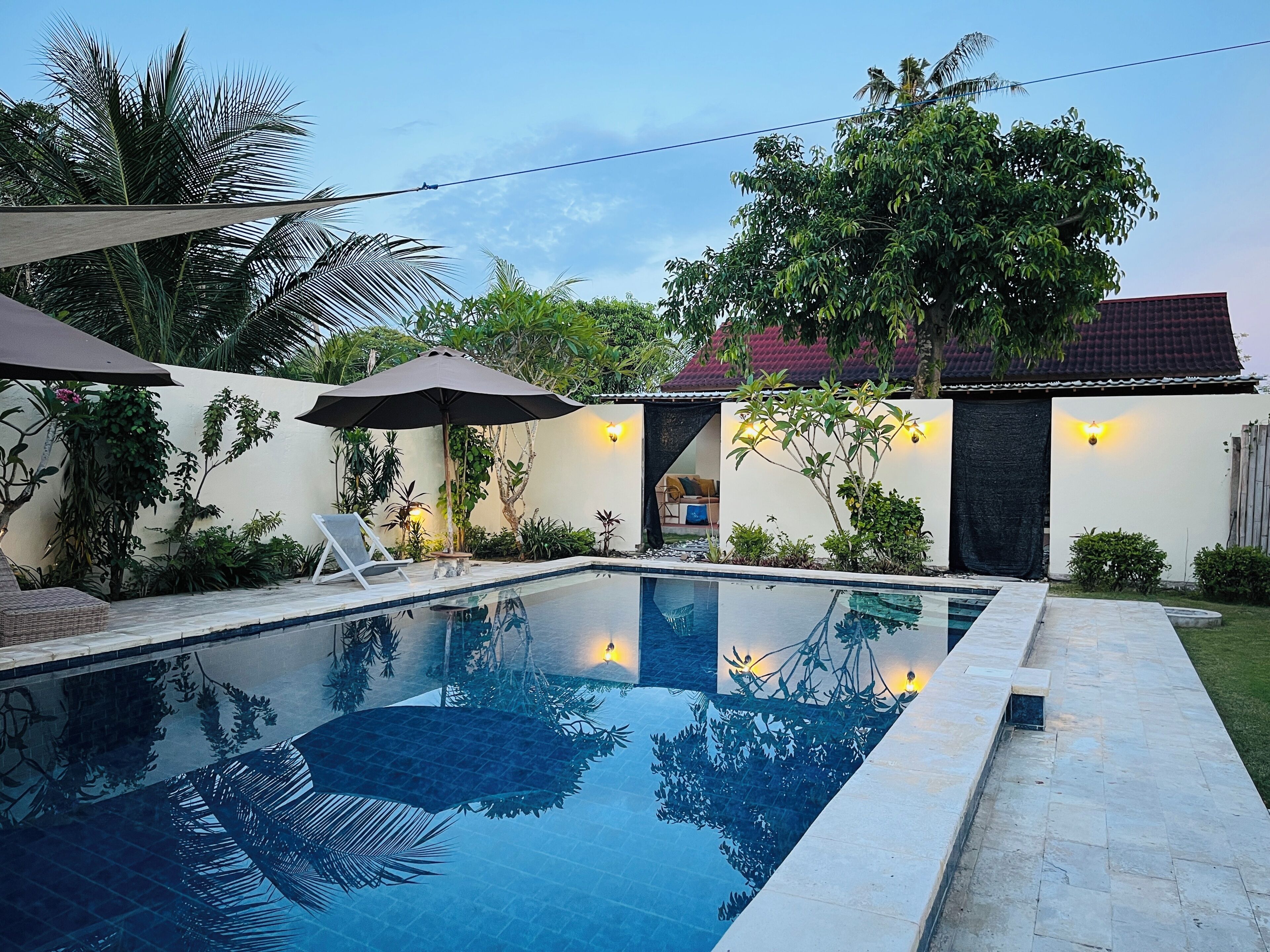 A swimming pool with a tiled deck, surrounded by lush greenery and a house with a tiled roof.