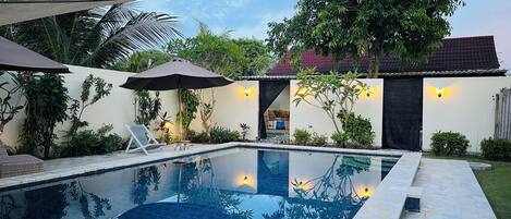 A swimming pool with a tiled deck, surrounded by lush greenery and a house with a tiled roof.