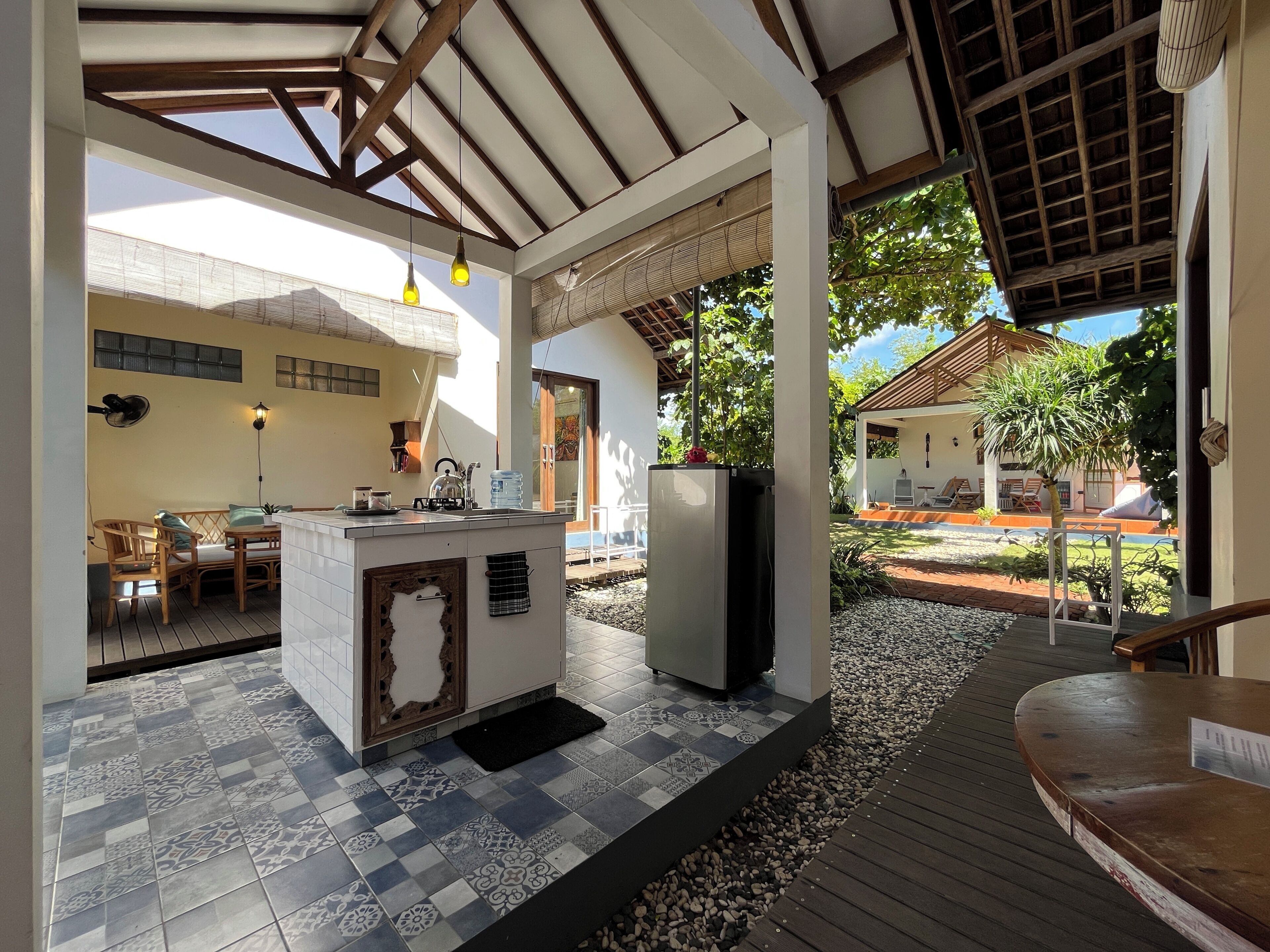 Un área de cocina al aire libre, techada, con piso de baldosas, una isla central con pileta y un refrigerador.