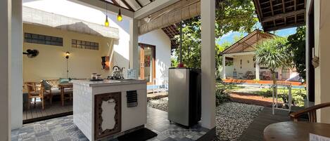 Un área de cocina al aire libre, techada, con piso de baldosas, una isla central con pileta y un refrigerador.
