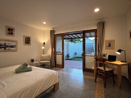 Un dormitorio con una cama, un escritorio, una silla, una computadora portátil y vista a un jardín por una puerta abierta.