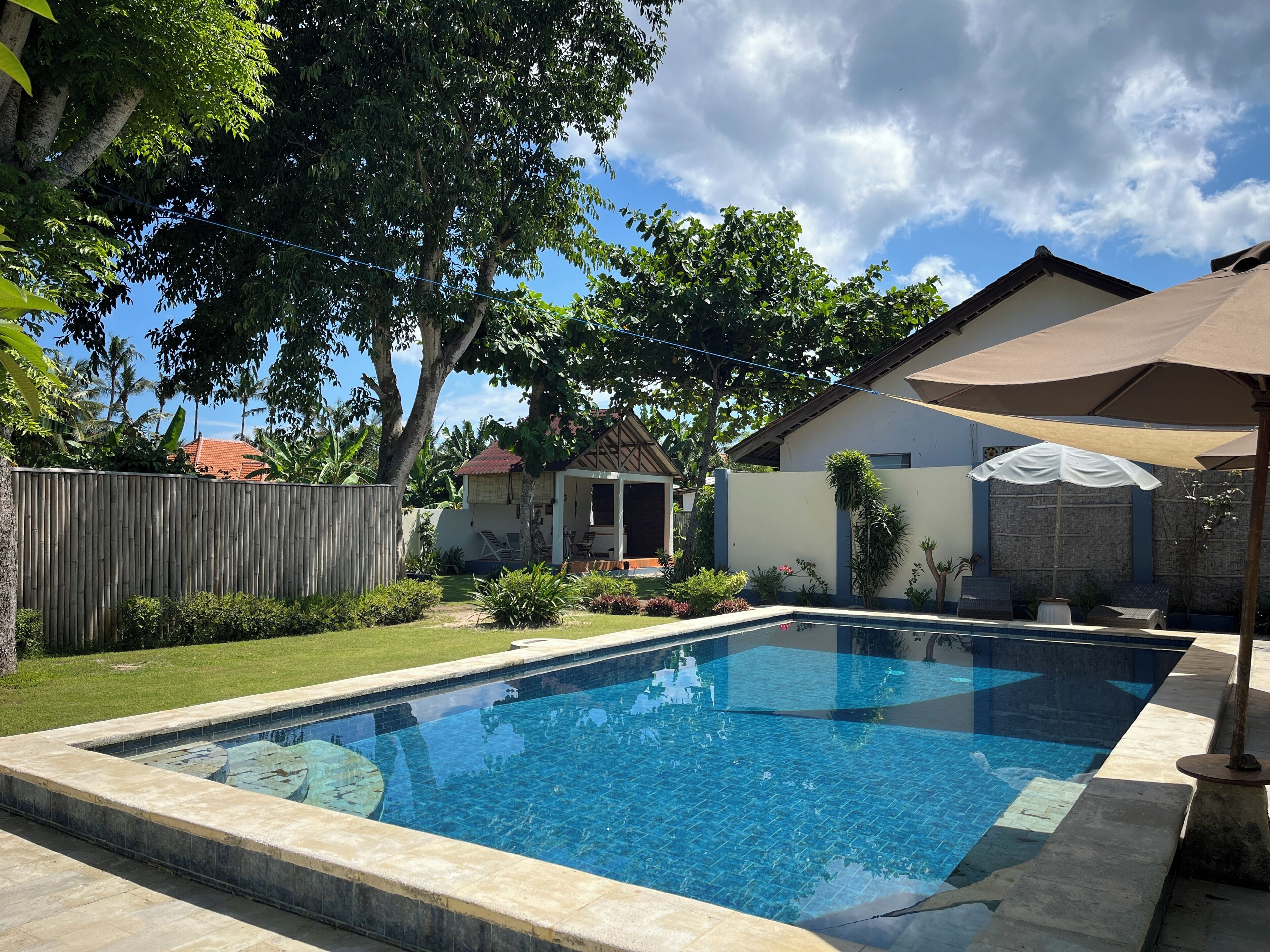 A swimming pool with a tiled deck, surrounded by a wooden fence, a house with a red roof, and a covered patio area.