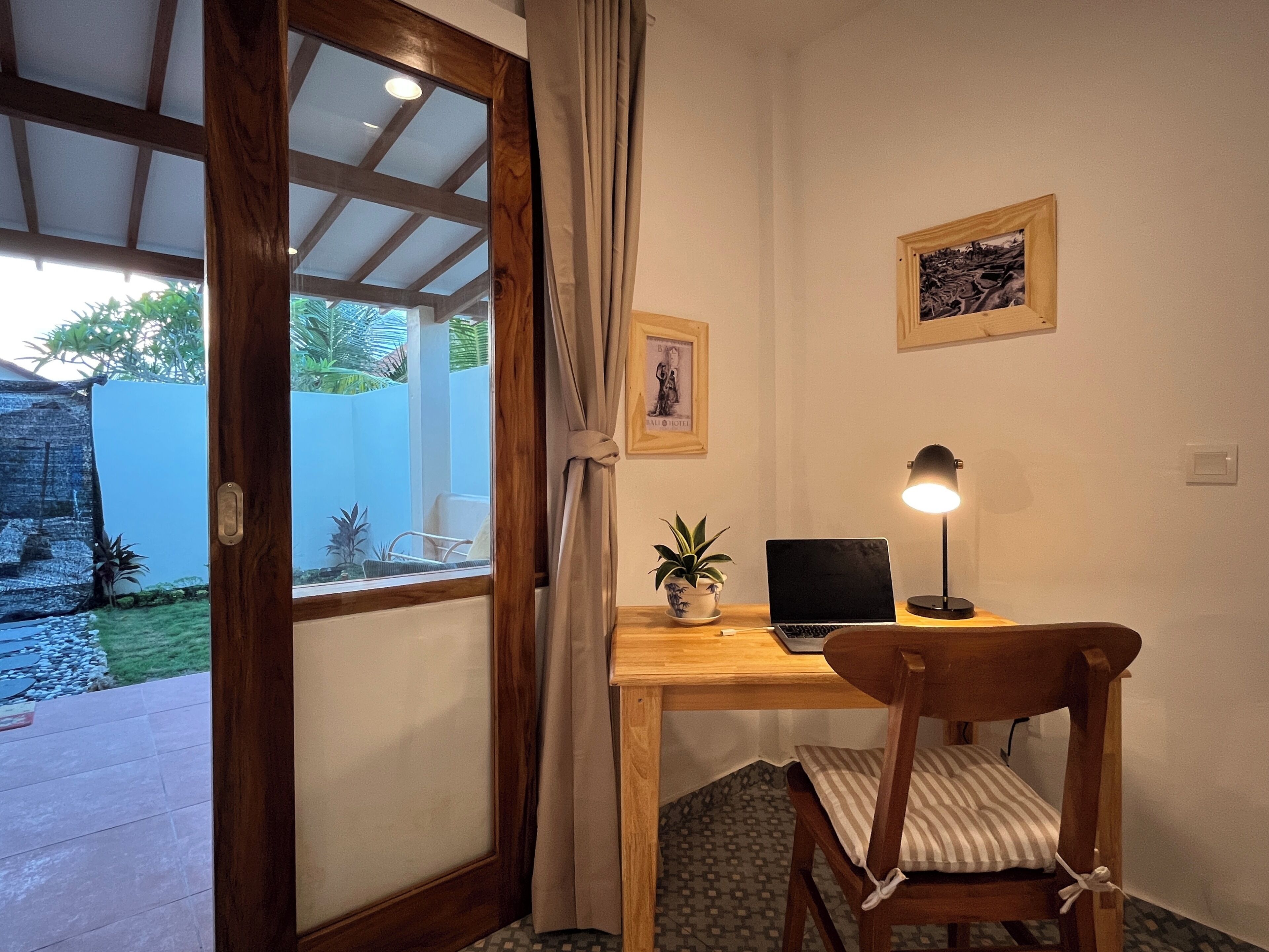 A room with a wooden desk, a laptop, a lamp, a chair, a potted plant, and a door leading to an outdoor area with a view of greenery.