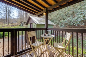 Um deck de madeira com duas cadeiras e uma mesa, com vista para uma casa verde e árvores.