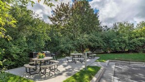 An outdoor patio area with multiple round tables and benches, surrounded by lush greenery and trees. - 1 King Bed | Non-Smoking, Kitchen (Milwaukee WI)