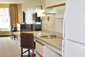 Una habitación de hotel compacta con kitchenette, una zona de comedor con mesa y sillas, un televisor de pantalla plana y un amplio ventanal con cortinas.