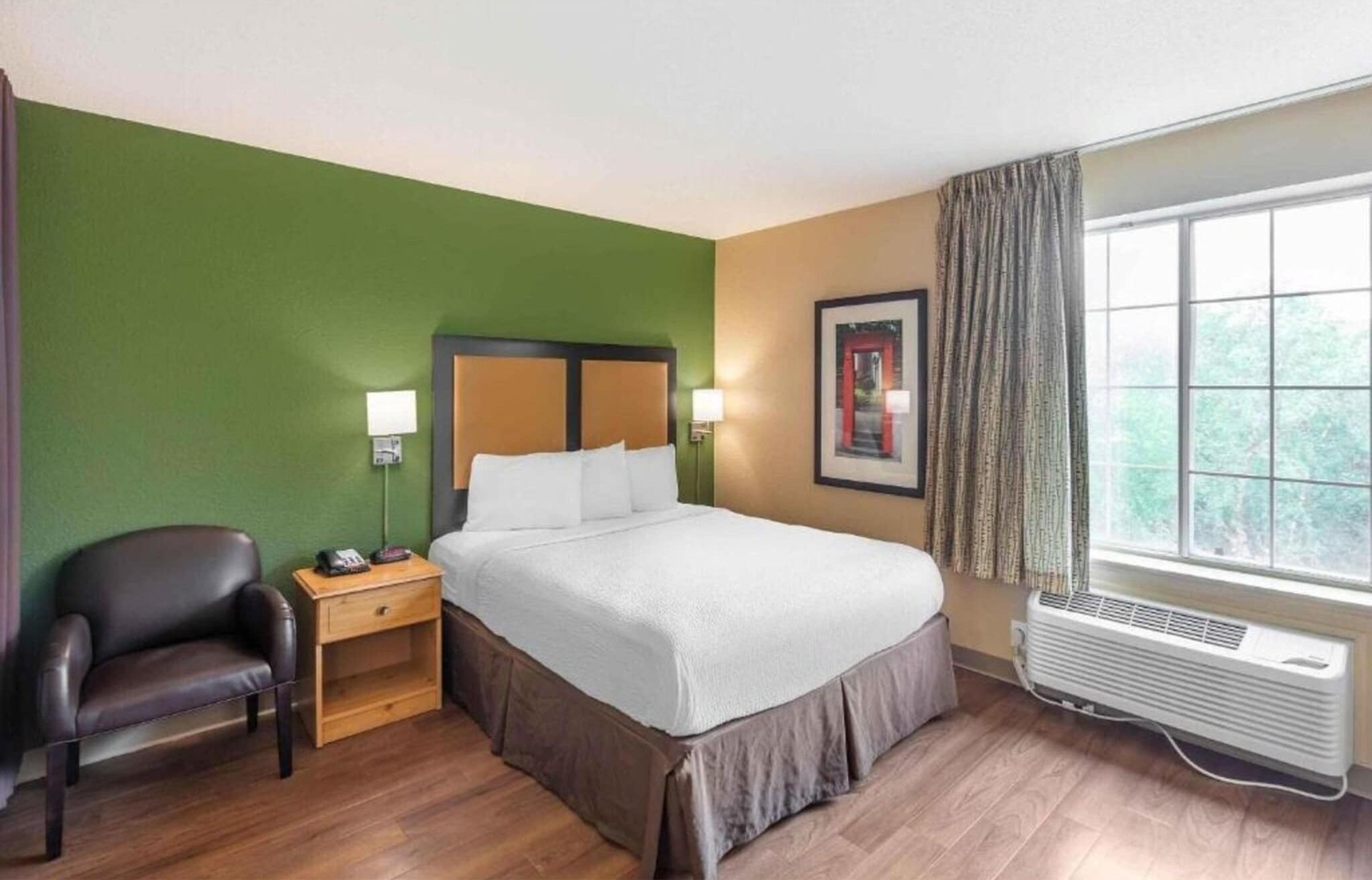 Hotelkamer met een bed, een stoel, een nachtkastje, een wandlamp, een lijst met een foto en een raam met gordijnen.