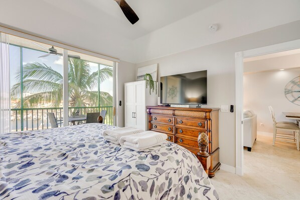 Un dormitorio con cama, cómoda, televisor y balcón con palmeras.
