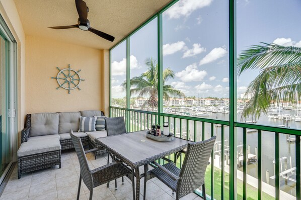 A balcony with wicker furniture, a table with a tray, and a view of a marina with boats and palm trees. - Sunsets & Sails! Waterfront Punta Gorda Condo (Punta Gorda)