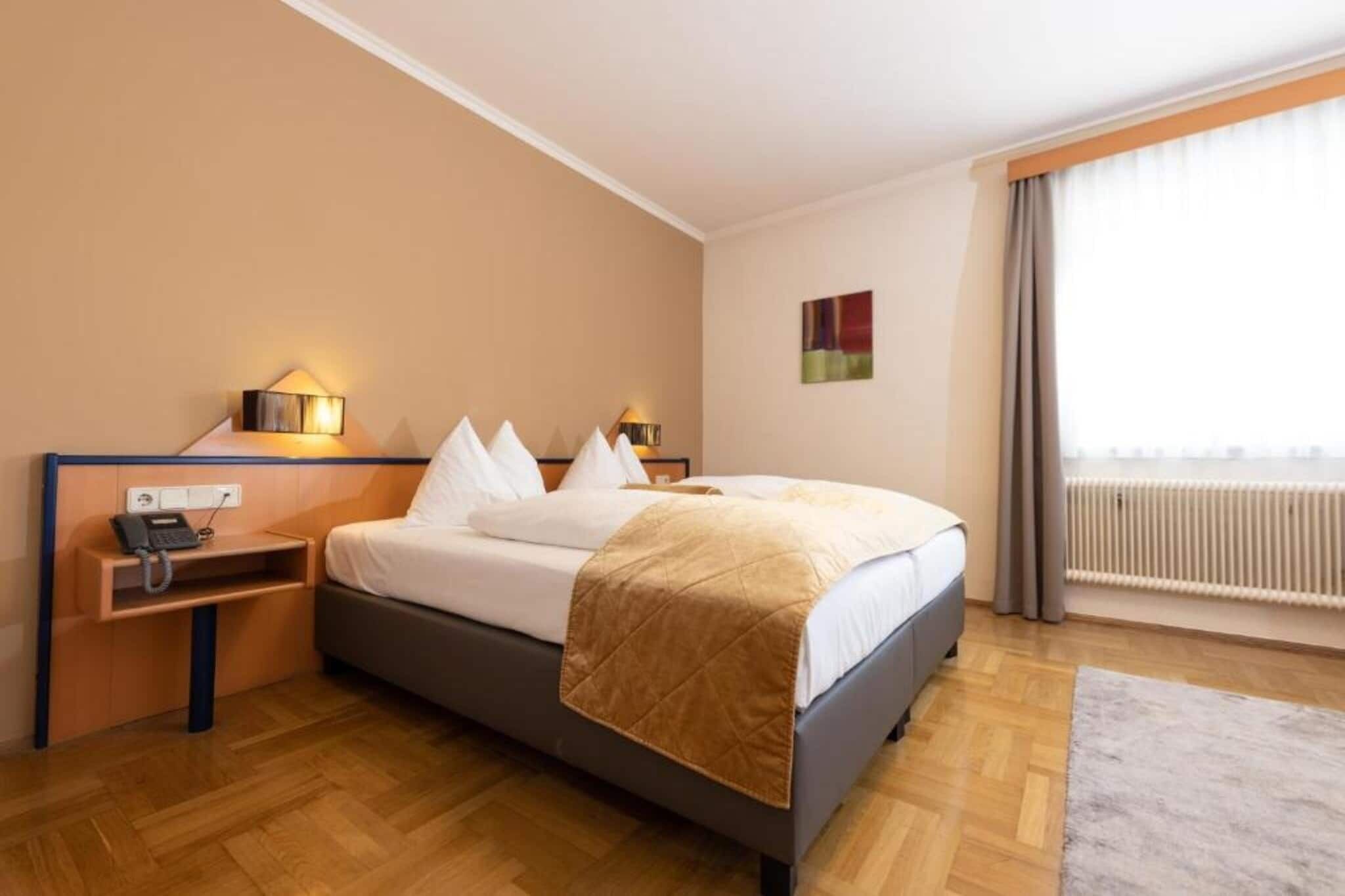 A hotel room with a bed, bedside tables, a telephone, a radiator, and a window with curtains.