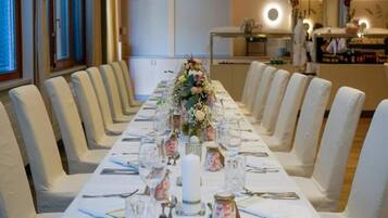 Una larga mesa de comedor dispuesta para un evento formal, con sillas a ambos lados, un centro de mesa floral y una cocina al fondo.