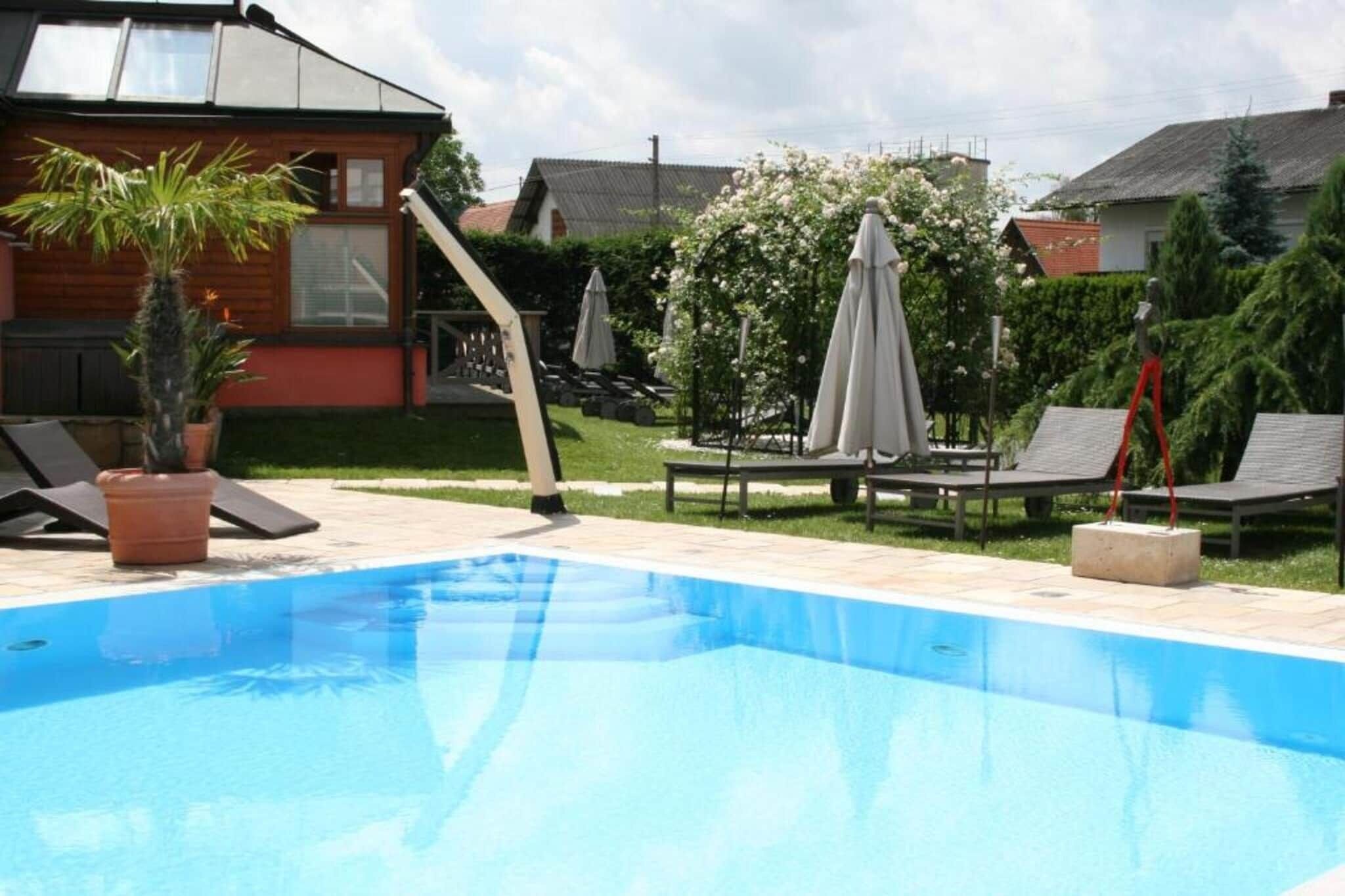 En swimmingpool med solstolar och parasoller, omgiven av en trädgård och ett trähus.