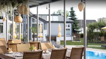 Un área de comedor al aire libre con muebles de mimbre, adornos colgantes y vistas a una piscina y edificios al fondo.