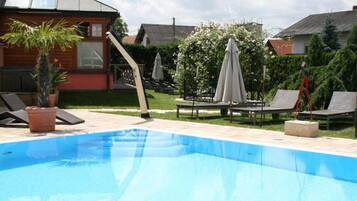 A swimming pool with lounge chairs and umbrellas, surrounded by a garden and a wooden house.