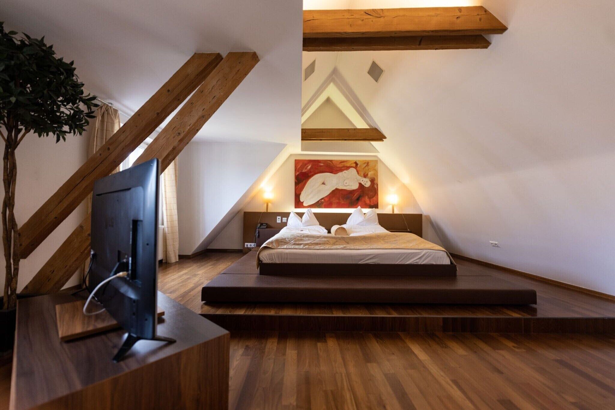 A bedroom with a sloped ceiling, a bed, a TV, and a painting.