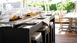 Un buffet avec des présentoirs en plastique transparent recouvrant les aliments, une table en osier et des sièges d’extérieur protégés par des parasols.