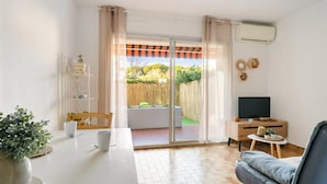 Un salon comprenant un canapé, un téléviseur, une table à manger avec des chaises, une porte-fenêtre coulissante donnant sur un jardin et une petite table d’appoint avec une plante.