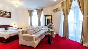 A hotel room with a bed, a sofa, a small table with a vase of flowers, and large windows with curtains. - Garni Hotel Royal Crown (Subotica)