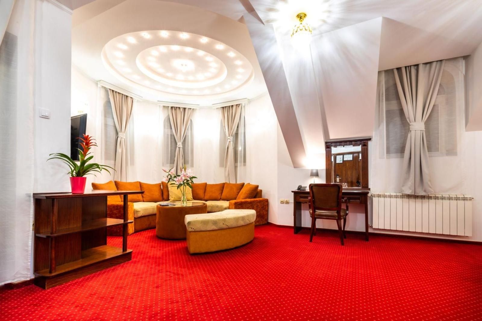 Habitación de hotel con alfombra roja, un sofá, una mesita con un florero, una silla y un escritorio con espejo.