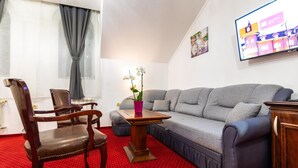 A living room with a grey sofa, wooden chairs, a red carpet, and a television on the wall. - Garni Hotel Royal Crown (Subotica)
