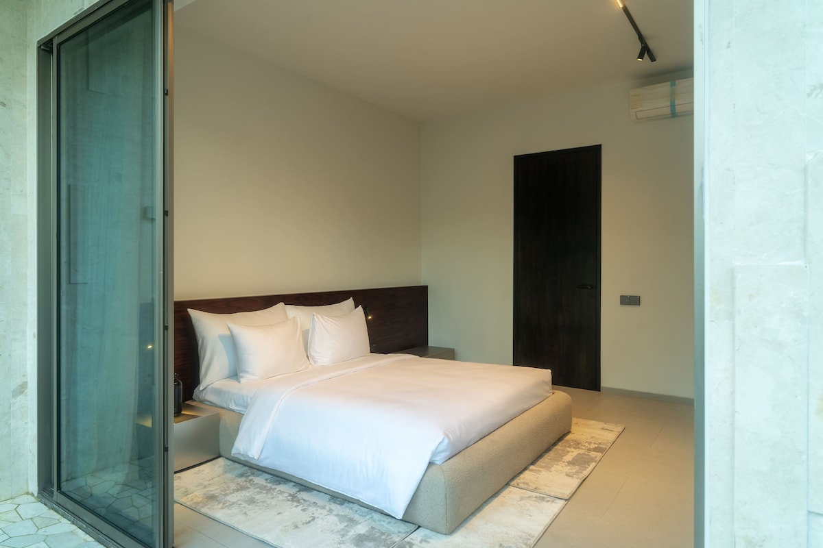 A bedroom with a bed, a nightstand, a wooden headboard, a sliding glass door, and a dark door.