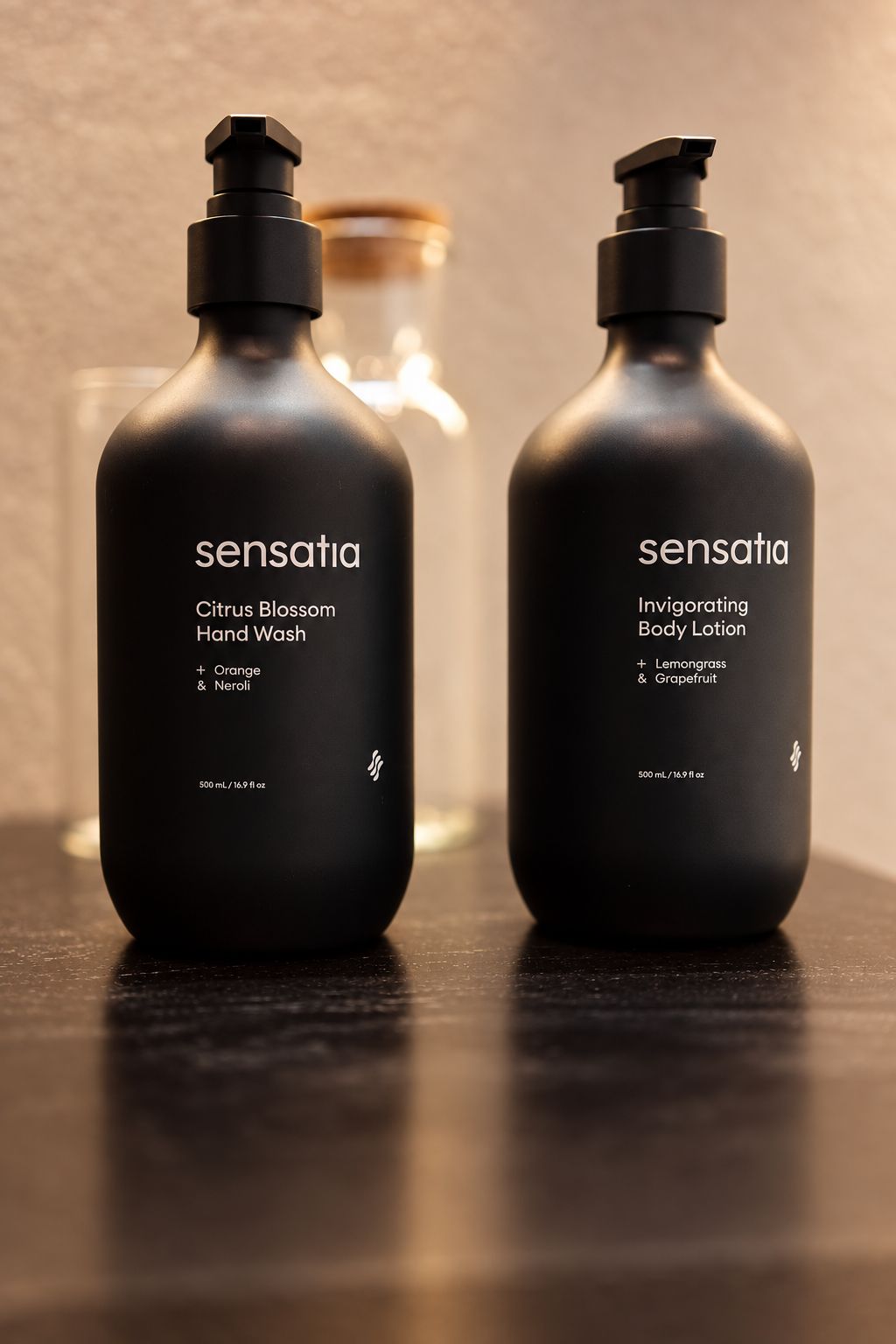 Two black bottles of Sensatia hand wash and body lotion on a dark surface with a blurred background.