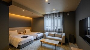 Moderni hotellihuone, jossa on kaksi sänkyä, sohva, sohvapöytä ja suuri ikkuna, jossa on sälekaihtimet.