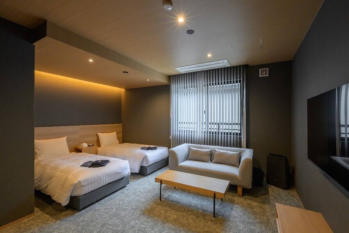 Ein modernes Hotelzimmer mit zwei Betten, einem Sofa, einem Couchtisch und einem großen Fenster mit Jalousien.