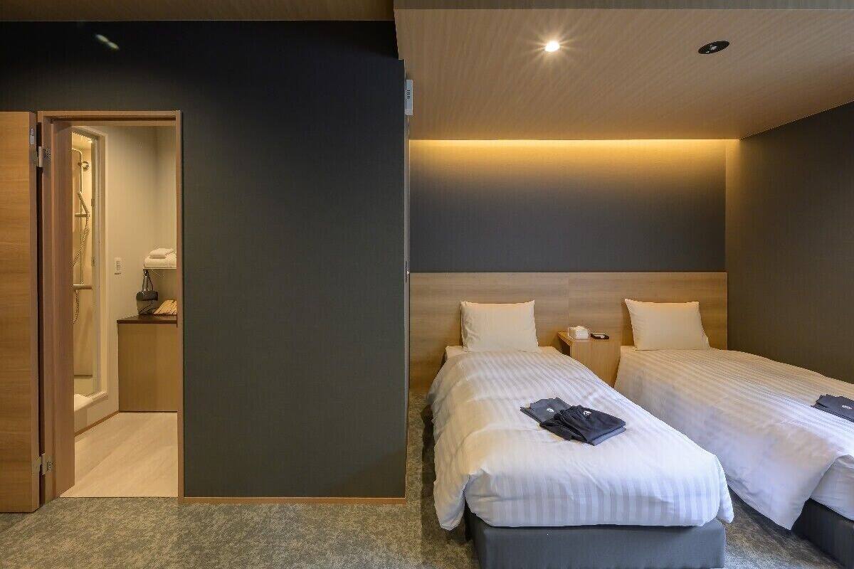 Ein Hotelzimmer mit zwei Betten, einer Holztür, einem kleinen Nachttisch mit Lampe und einem sichtbaren Badezimmer.
