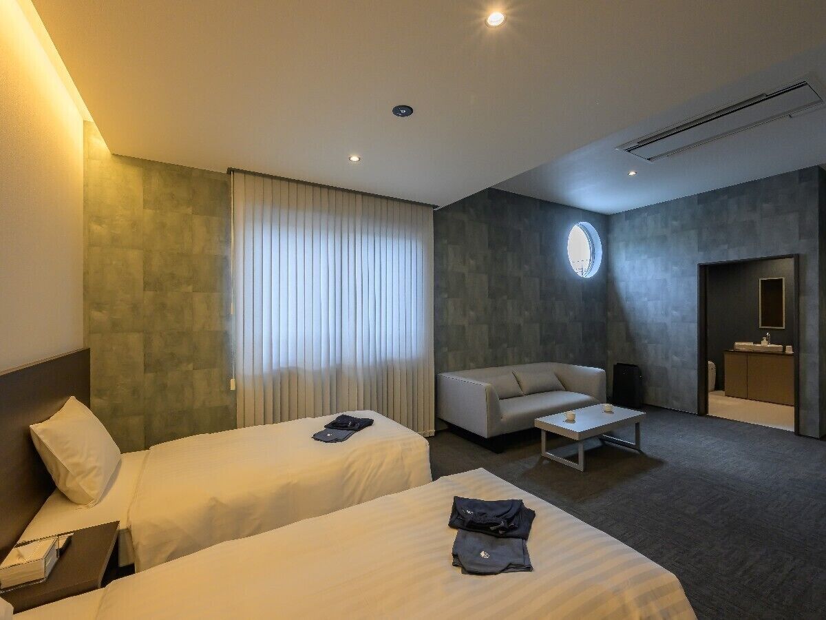 A hotel room with two beds, a sofa, a small table, a round mirror, and a bathroom visible through an open door.
