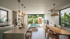 A modern kitchen with a central island, bar stools, a dining table, pendant lighting, and a view of a pool and greenery. - Villa Enola Uluwatu (Uluwatu)