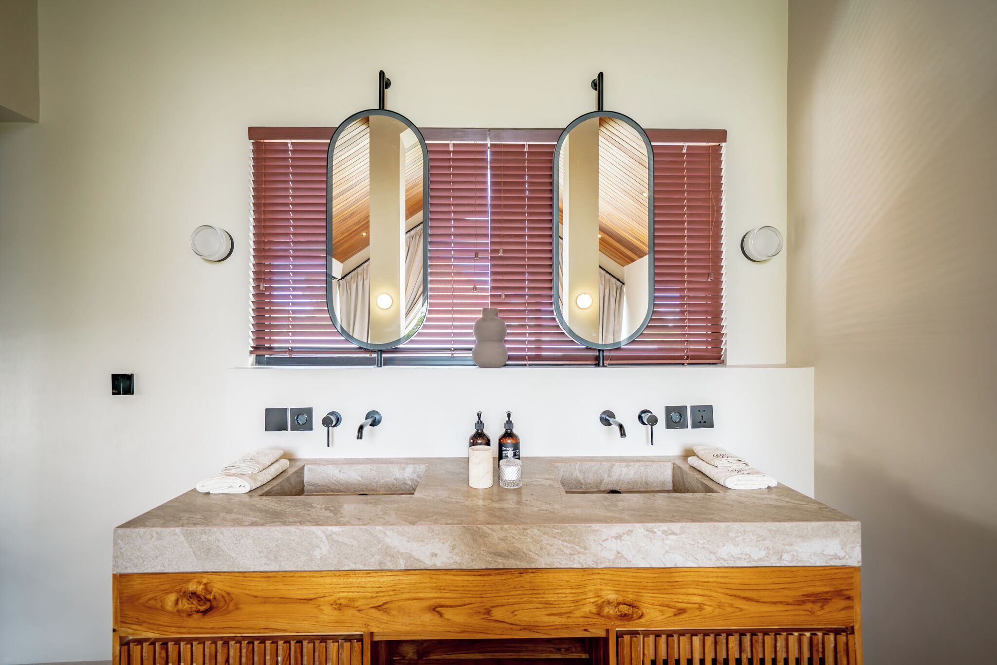 A bathroom with a double sink, two mirrors, a wooden vanity, and a window with blinds.