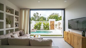 A modern living room with a sofa, a coffee table, a TV, and a view of a pool and garden. - Villa Enola Uluwatu (Uluwatu)