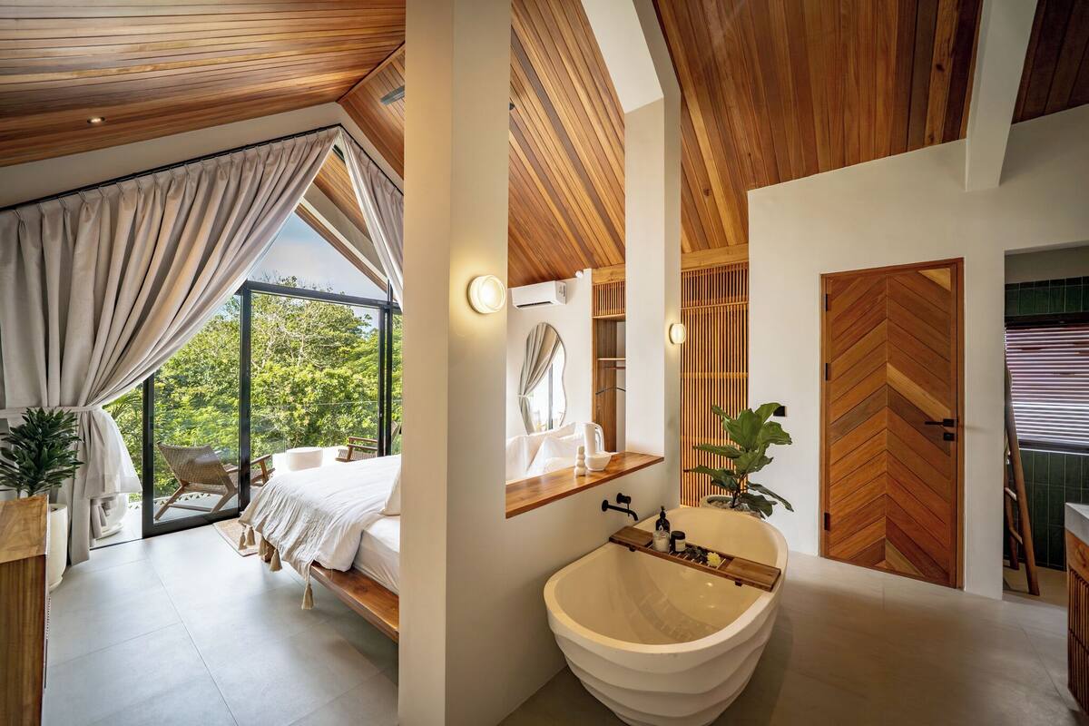 A modern bedroom with a large window, a wooden ceiling, a bed, and a view of greenery.