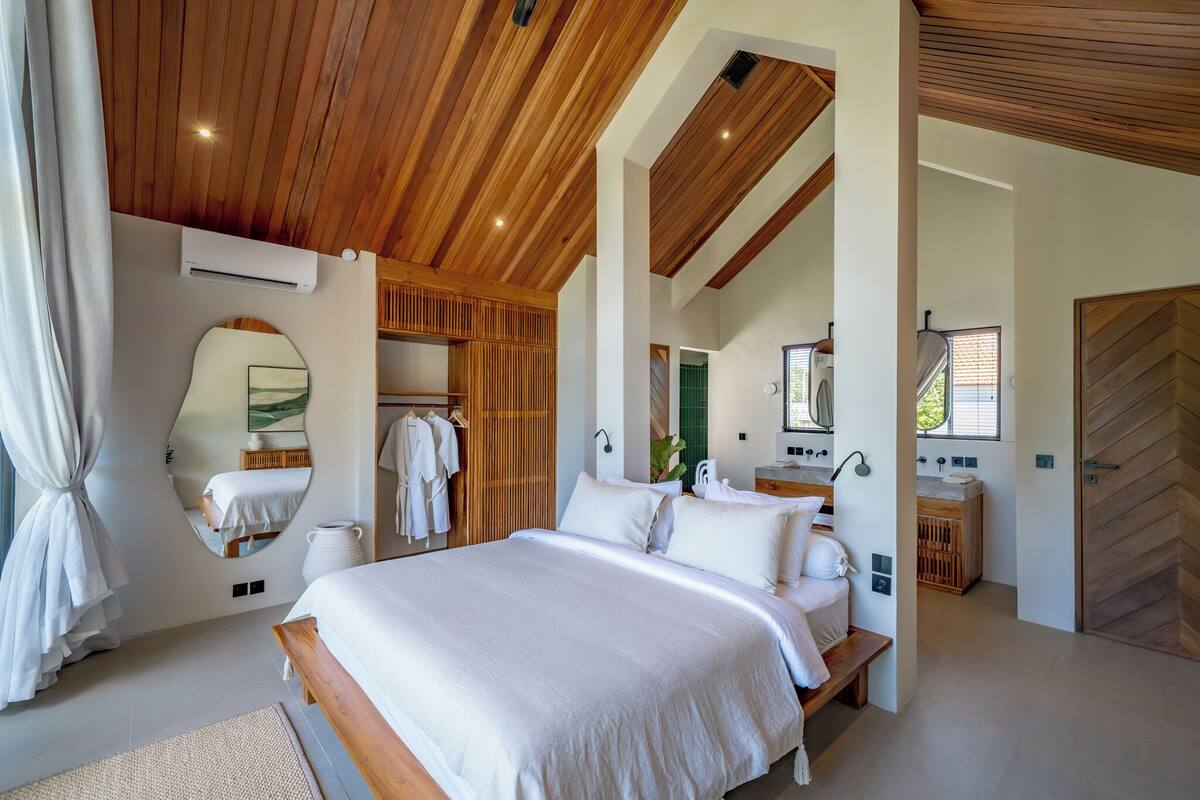 A bedroom with a wooden ceiling, a large bed, a walk-in closet, and a bathroom with a wooden herringbone door.
