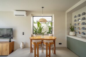 A modern dining area with a wooden table, chairs, a vase of flowers, and a large window with plants. - Villa Enola Uluwatu (Uluwatu)