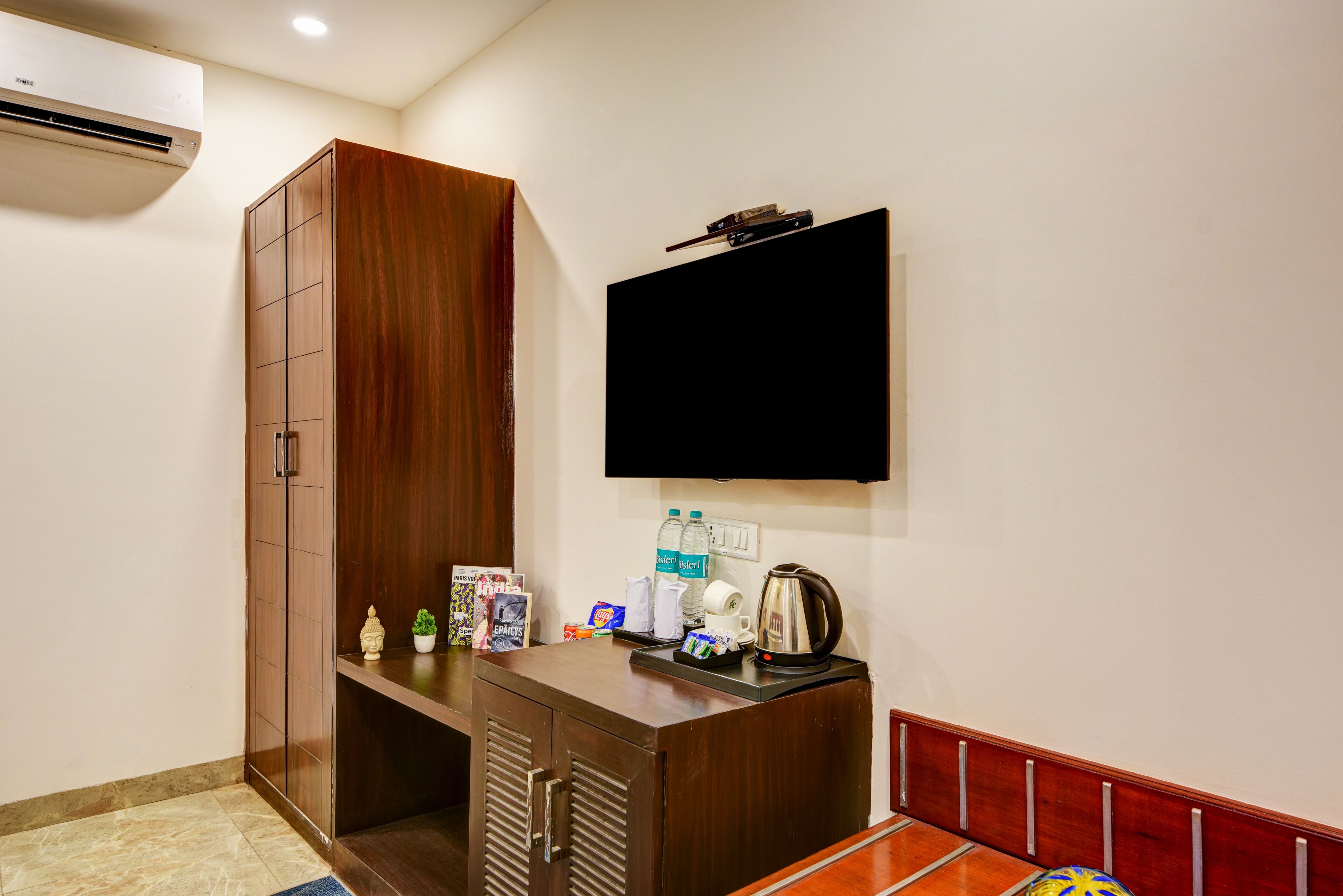 A hotel room with a wooden wardrobe, a flat-screen TV mounted on the wall, a small desk with a kettle, and a red cushioned bench.