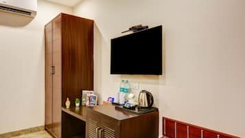 A hotel room with a wooden wardrobe, a flat-screen TV mounted on the wall, a small desk with a kettle, and a red cushioned bench.
