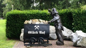 A statue of a man with a shovel and a cart labeled 'Glück Auf' in front of a hedge and rocks.