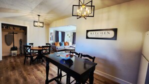 Uma sala de jantar com mesa de madeira, cadeiras e um candelabro. Há um aviso indicando cozinha na parede.