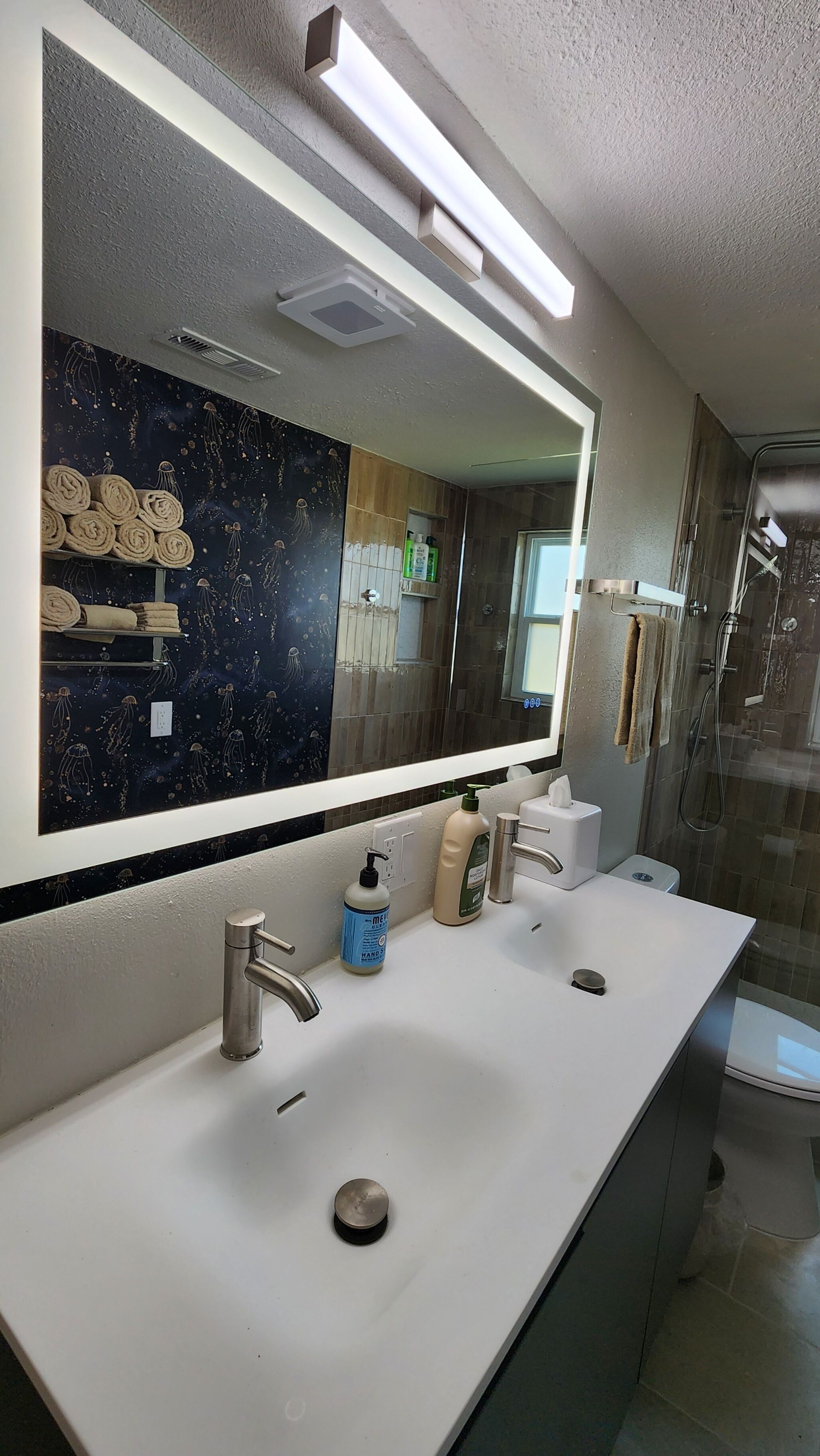 Une salle de bain dotée d’un grand miroir, d’un lavabo blanc, d’un distributeur de savon manuel et d’une cabine de douche en verre.